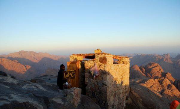 Small stone restroom labeled "W.C." stands on a rocky mountaintop at sunrise or sunset. A person is visible in front of the building, and extensive mountain ranges spread out in the background beneath a clear sky.