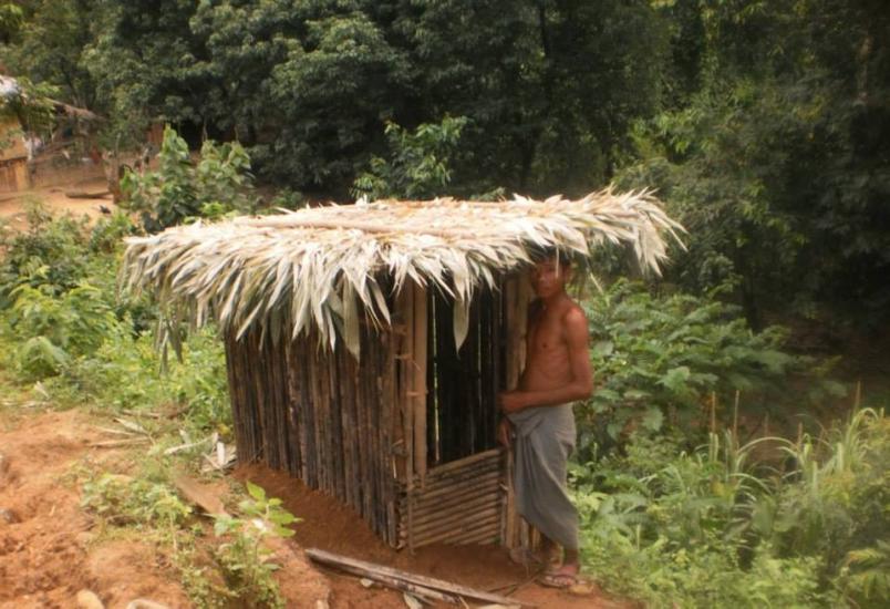 A person stands beside a small wooden structure with a thatched roof, surrounded by lush greenery and trees. The area appears rural and natural. The structure seems to be made from bamboo and other natural materials.