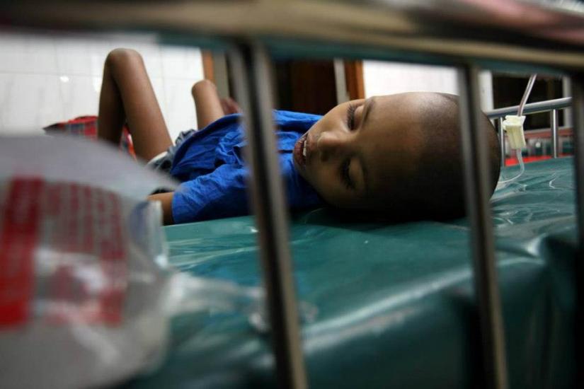 A child lies on a hospital bed, appearing ill and resting on their side. They are wearing a blue shirt, with an IV line visible. The setting suggests a medical or hospital environment.