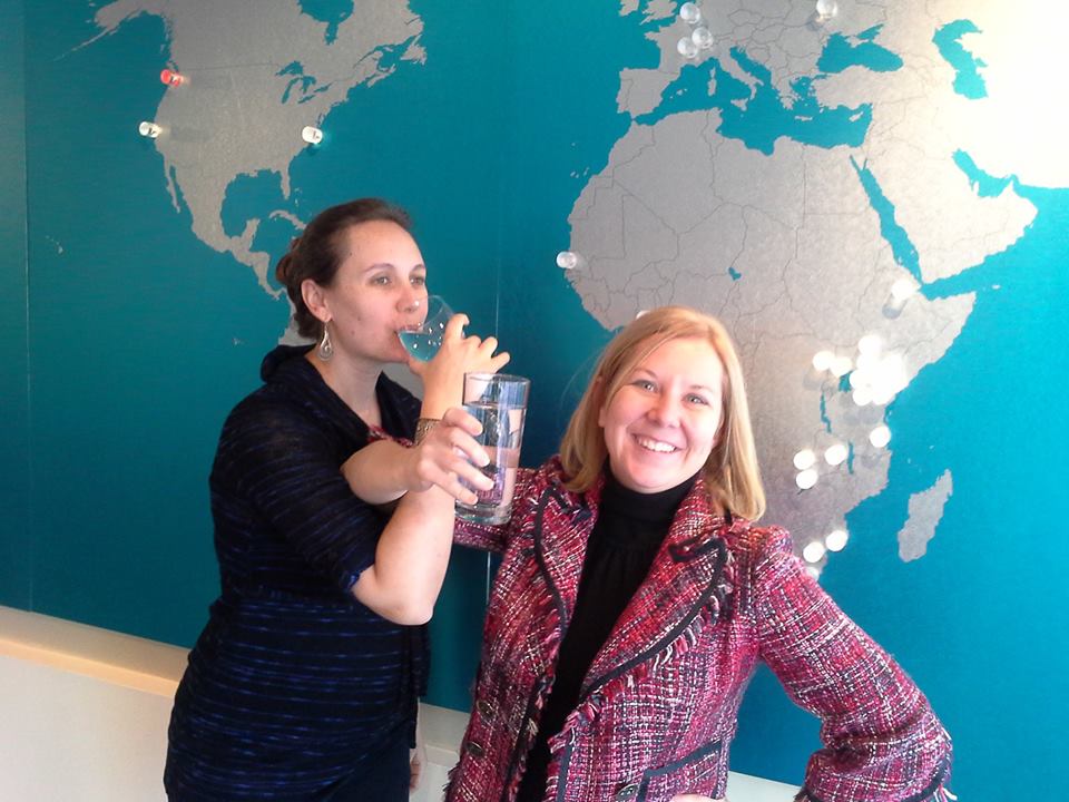 Two women are standing in front of a world map with pins marking various locations. The woman on the left, wearing a dark top, is drinking from a glass of water, while the woman on the right, wearing a red and black patterned jacket, is smiling at the camera.