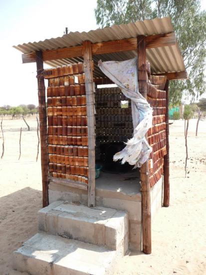 Image shows a small outdoor structure with walls made from rows of stacked plastic bottles. It has a corrugated metal roof and a tattered cloth hanging as a door. The structure is raised on a concrete platform with two steps leading up to it.