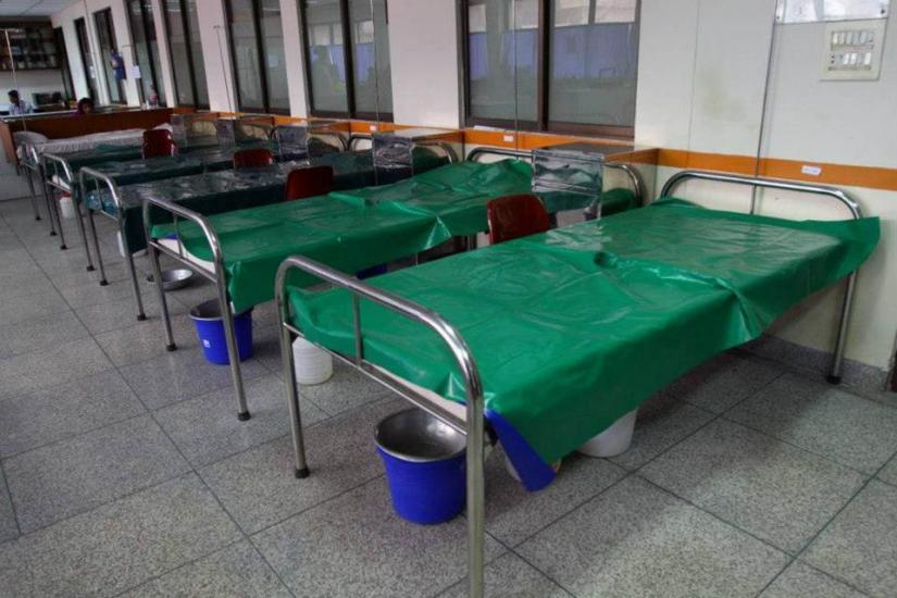 Rows of empty hospital beds with green covers, each accompanied by metal basins and blue buckets, situated in a hospital room with large windows and tiled floors.