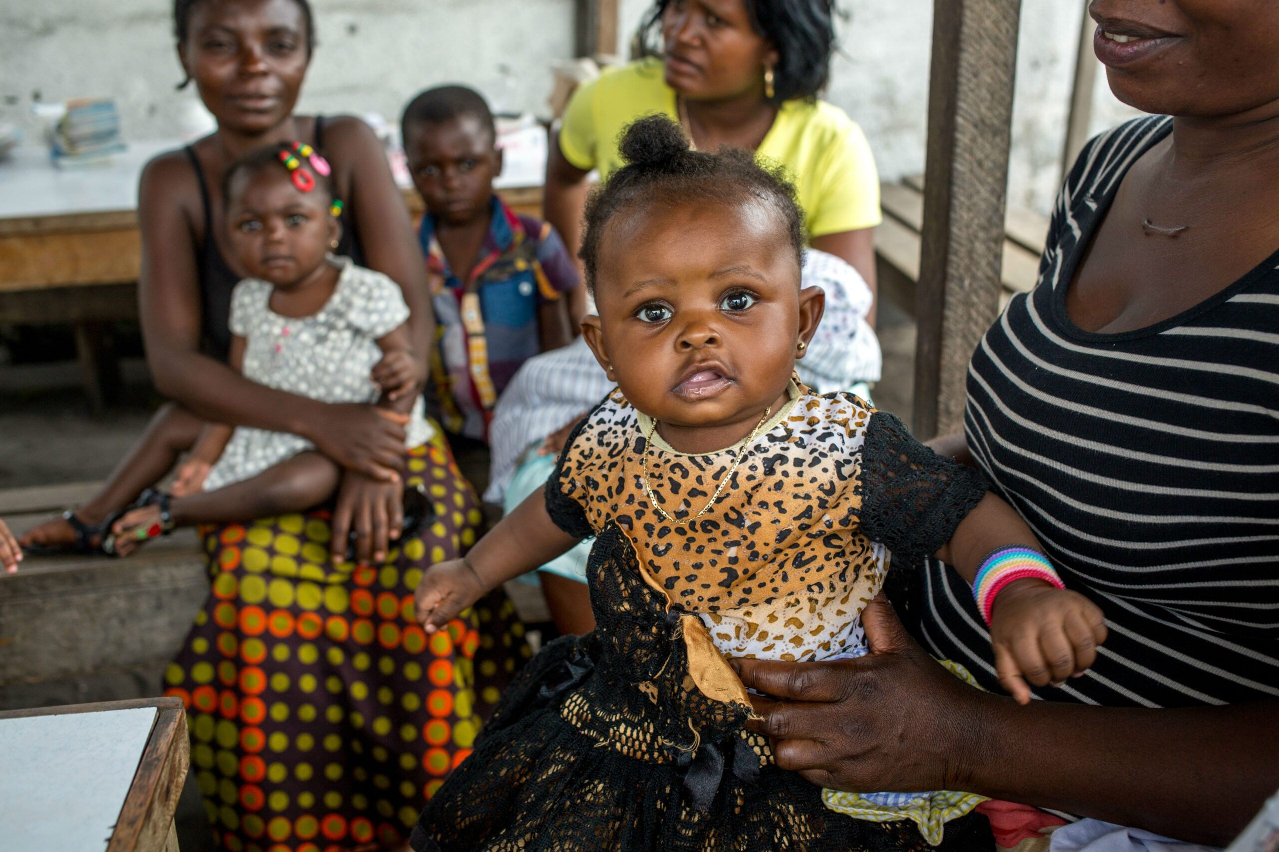 A group of women and children are gathered indoors. A woman is holding a baby with a ponytail, dressed in a leopard-print top and black skirt. Another woman, holding a baby, and two children are sitting in the background. The scene is casual and informal.