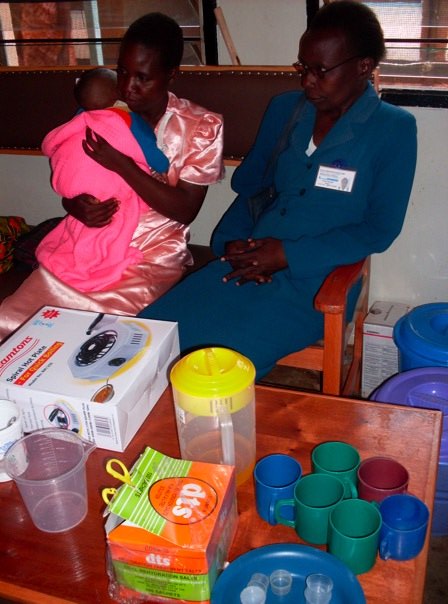 Two women sit on a wooden bench. The woman on the left is dressed in a pink outfit and is holding a baby wrapped in a pink blanket. The woman on the right wears a blue outfit and glasses. In front of them is a wooden table with various colorful containers and boxes.