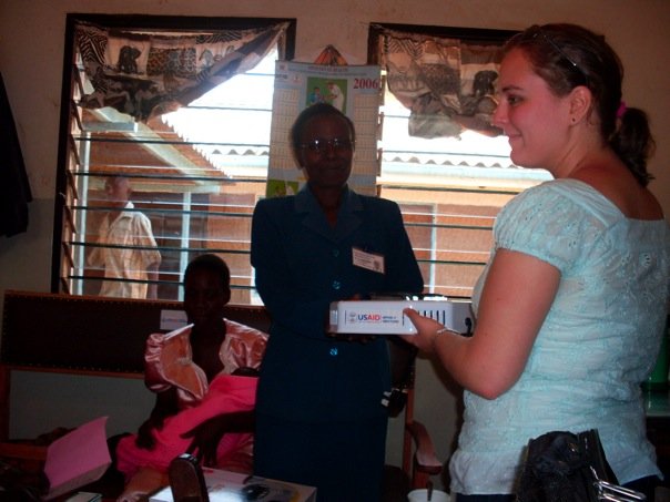 A woman in a light blue shirt hands a box to another woman in a business outfit. A young girl sits nearby wearing a pink dress. The scene appears to take place inside a room with barred windows and a calendar hanging on the wall.