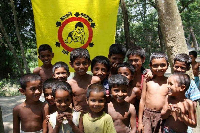 A group of smiling children stand together outdoors in front of a yellow banner with a logo featuring a monkey. The children, many of whom are shirtless, appear joyful and are standing in a wooded area with trees in the background.