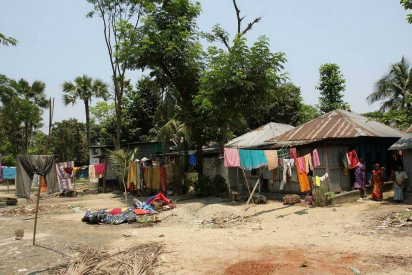 A rural scene with small houses featuring tin roofs. Clothing hangs on lines between trees. Several people stand near the houses, surrounded by trees and open sky. The ground is sandy with scattered items.