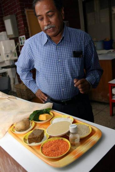 A person with a mustache and gray hair is standing and looking down at a tray filled with various foods, including lentils, a potato, spinach, and other items, in a kitchen or laboratory setting. The individual is wearing a blue checkered shirt with a pen in the front pocket.