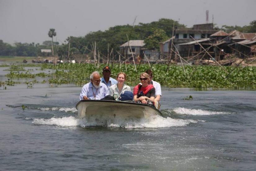 A small boat with four people navigates through a waterway surrounded by greenery and stilted structures. The passengers are wearing life jackets, and the background includes vegetation and buildings. The sky is clear and sunny.