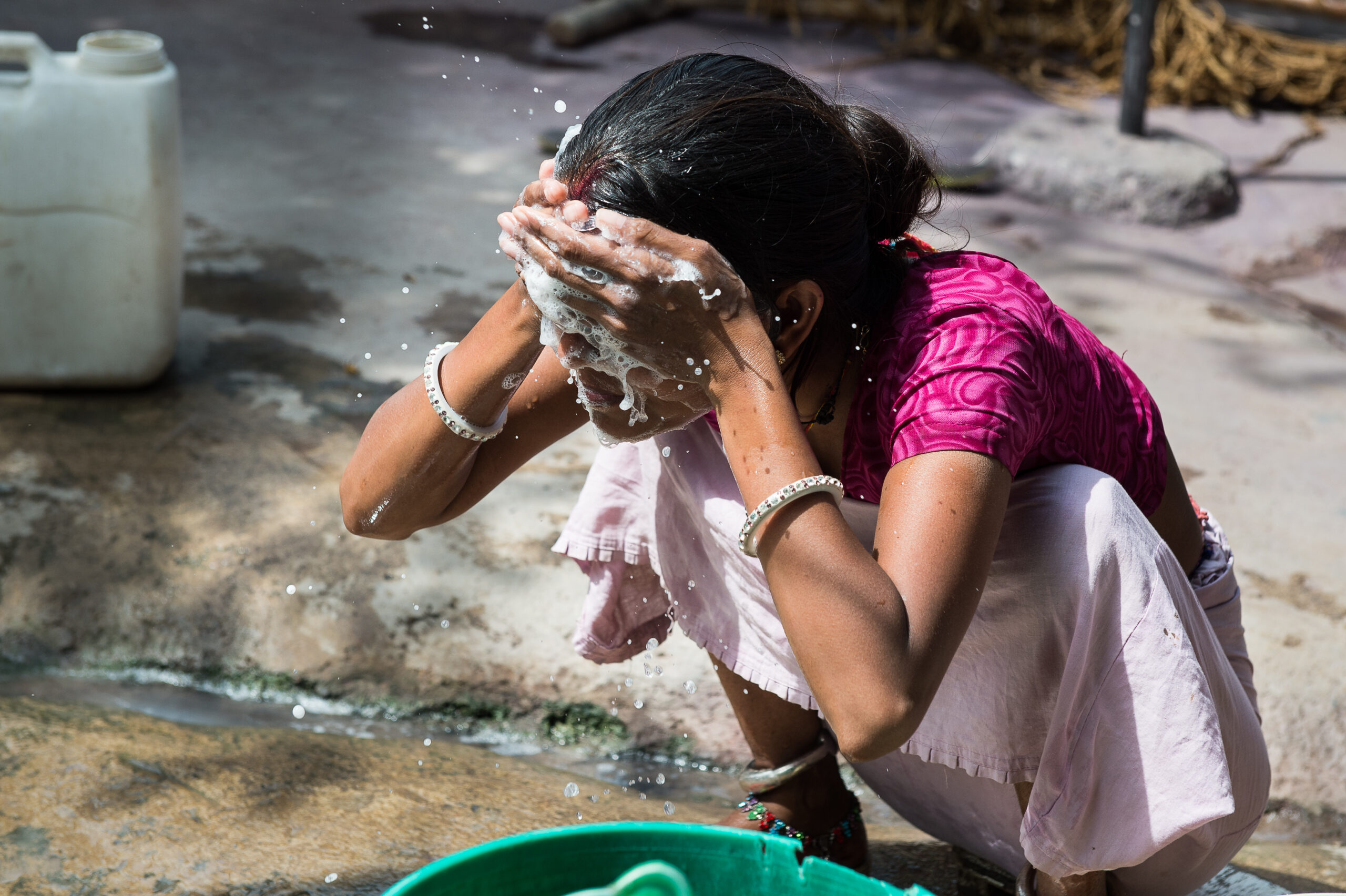 A person wearing a pink shirt and light pants squats near a green container, washing their face with soapy water. Water splashes around as they rinse their face. A white water container is visible in the background on the ground.