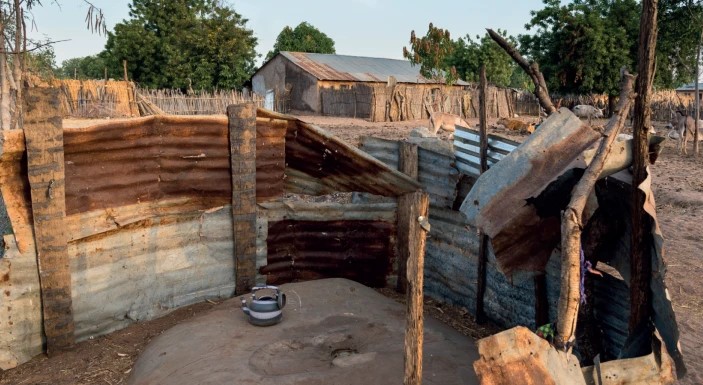 An outdoor area fenced with rusted metal sheets and wooden posts creates a small enclosure. Inside, a large clay cooking stove with a metal teapot sits in the middle. In the background, simple buildings and trees are visible under a clear sky.