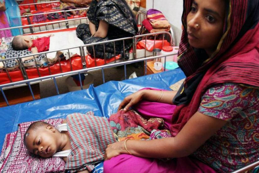 A woman dressed in colorful clothing sits on a blue mat beside a baby sleeping on a patterned blanket inside a metal crib. In the background, other women and babies are seen in similar settings, indicating a communal care or medical facility.