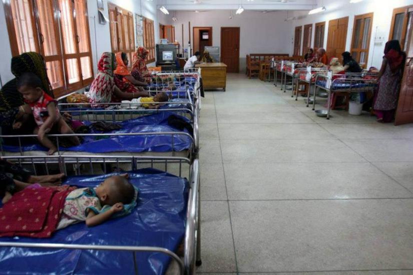 A room with several hospital beds, each holding a child, attended by women sitting beside them. The room has a long, open layout with a white floor, light walls, and large windows on one side. The atmosphere is somber and clinical.