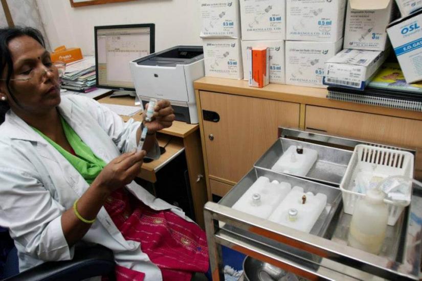 A woman wearing a lab coat sits in an office, holding a vial and a syringe. She is next to a medical cart with various supplies. Behind her are boxes of medical materials, a computer, and other office equipment.