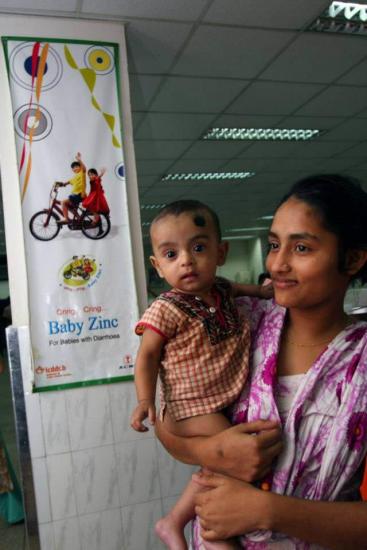 A woman is holding a baby in front of a banner advertising "Baby Zinc." The baby has a black dot on its forehead and is wearing a patterned outfit. The background shows indoor fluorescent lighting and part of a tiled wall. Both appear to be in a public space.