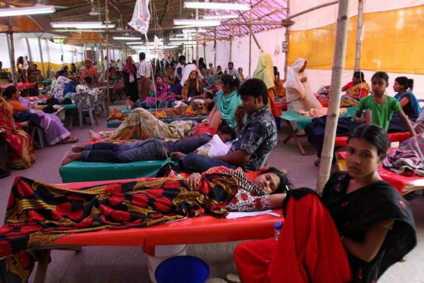 A large, crowded medical camp with numerous patients lying on cots. Women, men, and children are covered with blankets, some sitting and some lying down. Medical staff attend to the patients. The setting appears to be temporary, with makeshift arrangements and tarpaulin partitions.