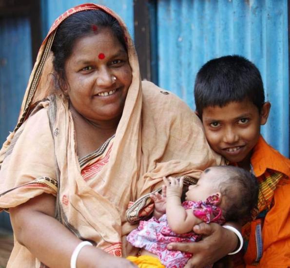 A woman in traditional attire smiles while holding a baby dressed in pink. A young boy in an orange shirt stands beside her, smiling. They are in front of a blue corrugated metal wall.