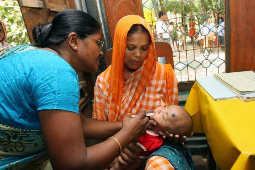 A healthcare worker administers an oral vaccine to a baby held by a woman in an orange headscarf, seated indoors near a window with decorative bars.