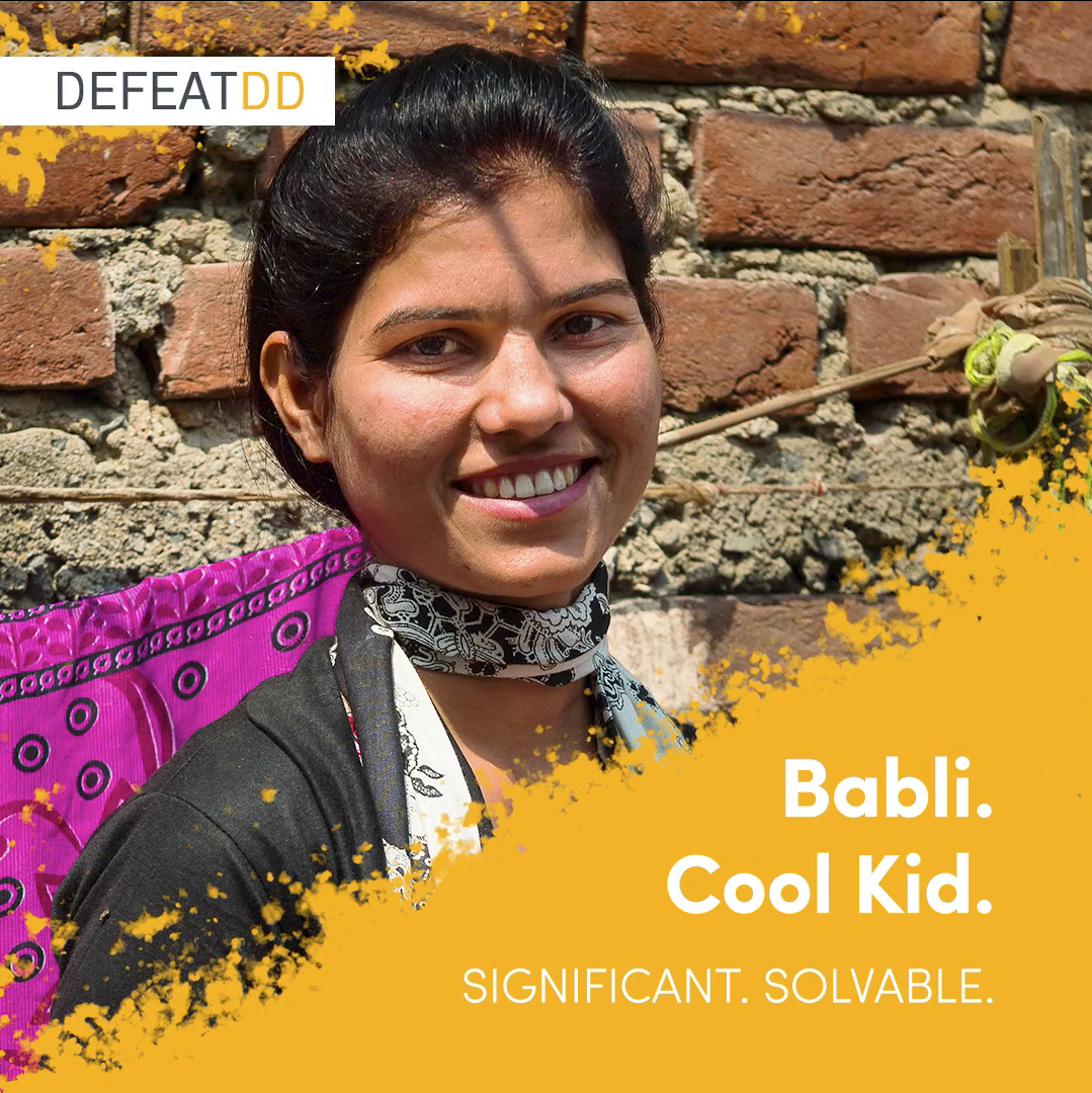 A young woman smiles warmly at the camera, standing in front of a brick wall with vibrant cloth hanging behind her. The image has text overlay: "Babli. Cool Kid. Significant. Solvable." The DefeatDD logo is in the top left corner.