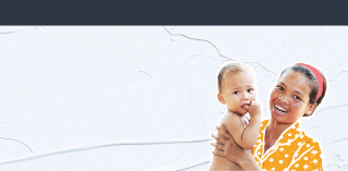 A smiling woman in an orange polka dot dress holds a baby. The baby has a curious expression and their finger in their mouth. The background is a plain textured wall.