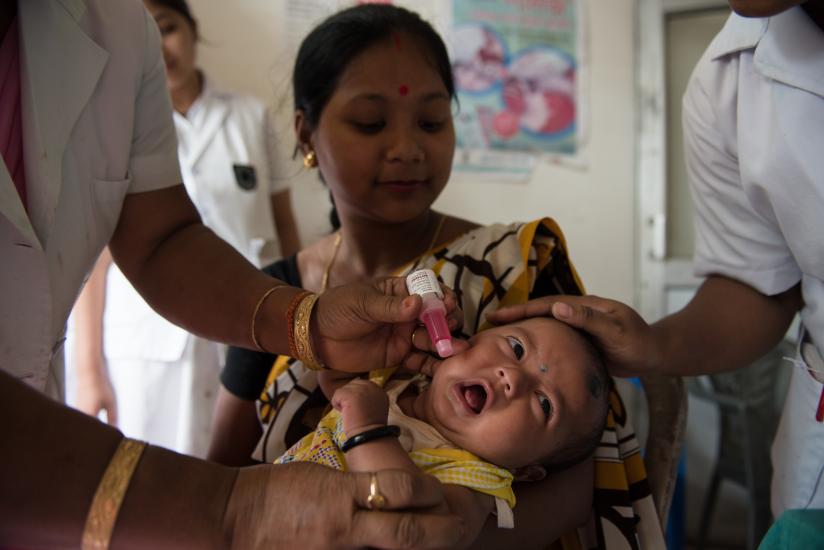 A healthcare worker administers an oral vaccine to a baby held by a woman in a clinic. Another healthcare worker supports the baby's head. The scene is set in a medical facility, with posters on the wall and a focus on the baby's open mouth.