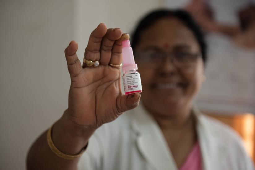 A person in a white lab coat holds up a small bottle of medication with a pink cap and label. The out-of-focus background contains indistinct medical posters. The person's face is slightly blurred, but they are smiling.