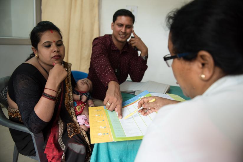 A mother and father are consulting with a health professional. The mother holds a sleeping baby, while the father points at a document on the table. The health professional, seen from behind, appears to be explaining something using the document.