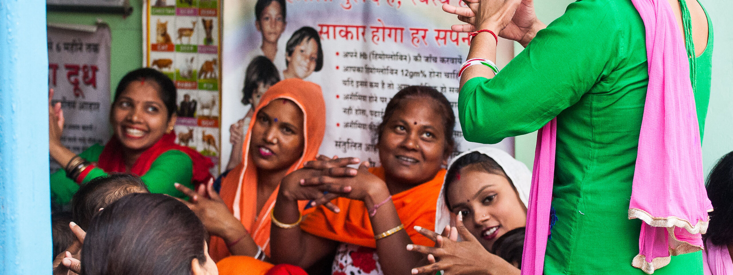 A group of women, dressed in colorful traditional clothing, sit closely together, smiling and clapping. One woman stands at the front, gesturing with her hands as if explaining something. Posters with writing and images are visible on the wall behind them.