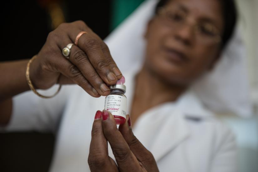 A healthcare professional in a white uniform and glasses holds a vial of a vaccine labeled "ROTAVAC." The person carefully adjusts the cap while the vial is held between their fingers, focusing attentively on the preparation process.