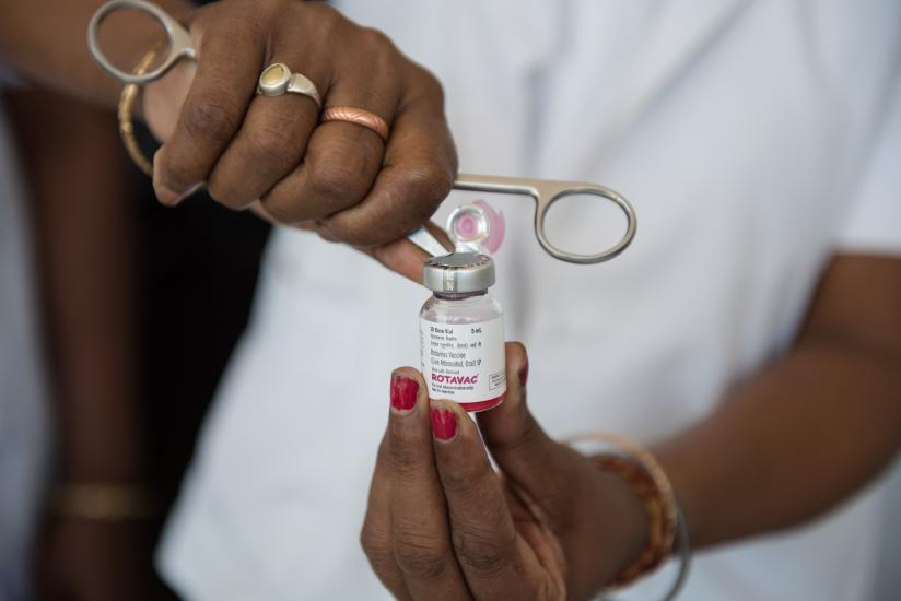 A person's hands are seen holding a vial labeled "ROTAVAC" with a pair of forceps. They are wearing a white coat and some rings. The vial cap is being removed.