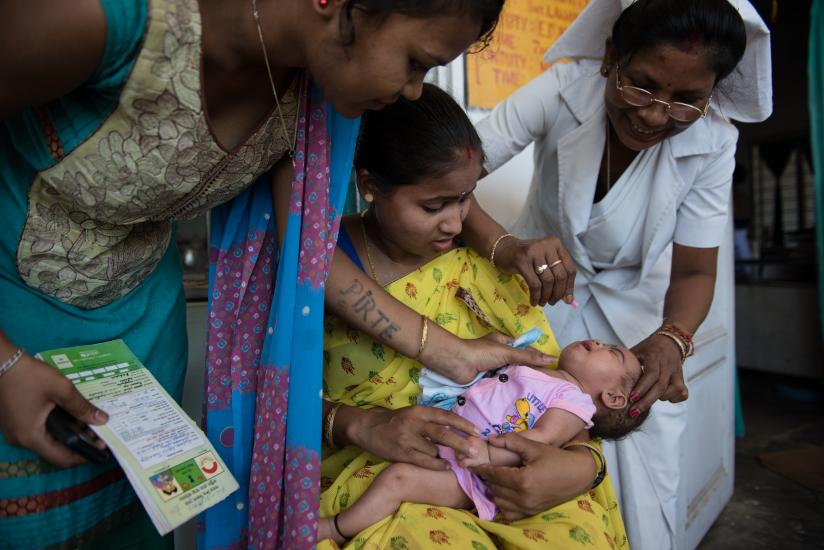 A healthcare worker administers a vaccine to a baby held by a seated woman in a yellow sari, while two other women stand by assisting and observing. The scene takes place in a medical setting, with the focus on the baby and the collaborative effort of the women.