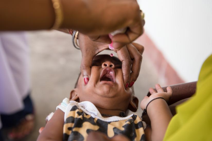 A child is receiving an oral vaccine administered by an adult. The child's mouth is being carefully held open, and the vaccine is being dropped inside using a dropper. The child is wearing a camouflage-patterned shirt.