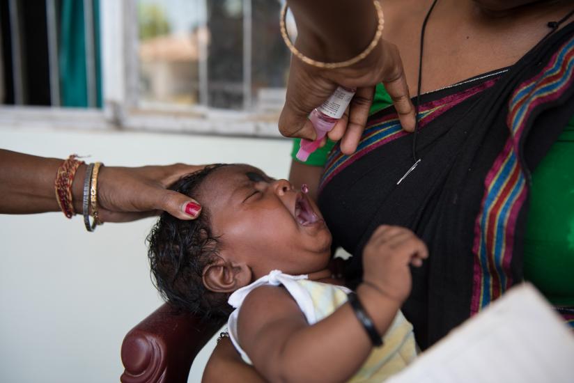 A crying infant sits on a woman's lap while receiving an oral vaccine from a healthcare worker inside a building. Another hand gently holds the baby's head for support during the vaccination.