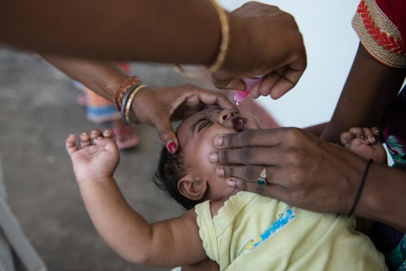 An infant, held gently by two adults, is receiving an oral liquid vaccine. The scene captures a close-up of caring hands administering the drops as part of a health initiative. The baby is wearing a light yellow garment.