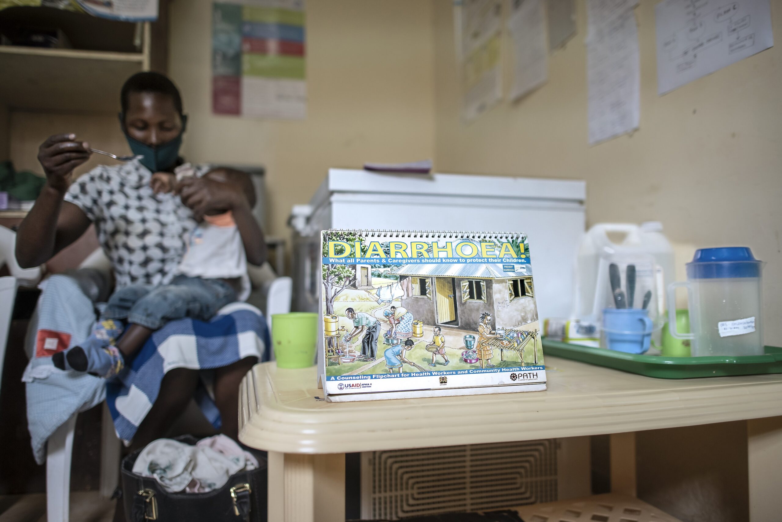 A person feeds a child in a room with a health poster titled "Diarrhoea" on a table in the foreground. Medical supplies are visible in the background.