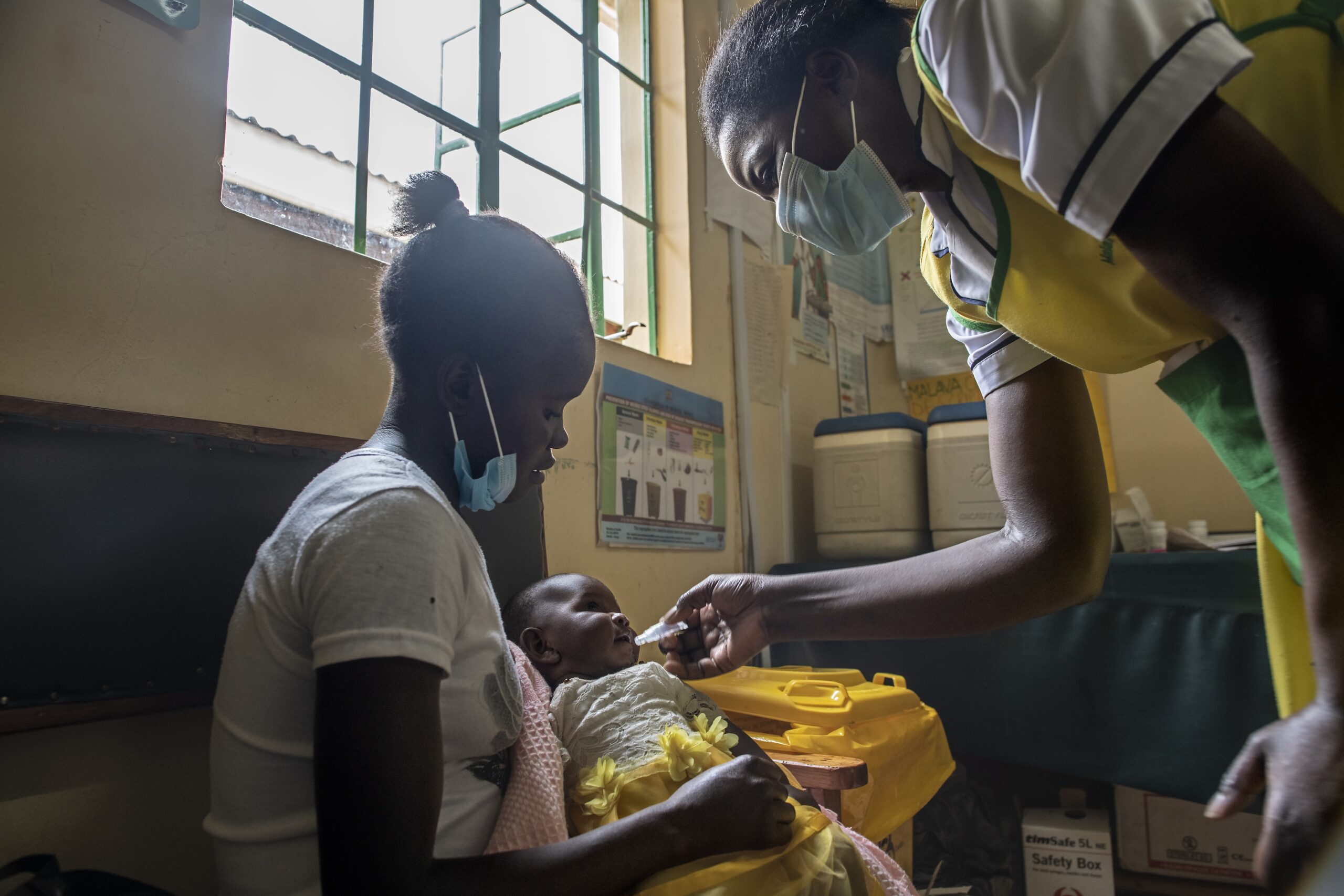 A nurse wearing a mask administers an oral vaccine to a baby held by a masked woman sitting on a chair. The setting appears to be a clinic, with medical supplies and posters in the background.