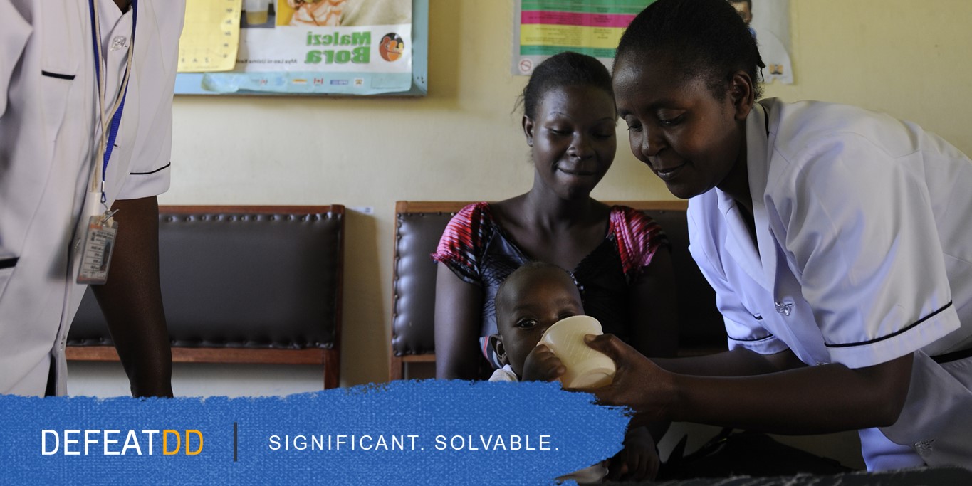 A healthcare worker in a white uniform helps a woman care for a small child sitting on her lap. The healthcare worker is holding a cup to the child's mouth. Text reads, "DEFEATDD. SIGNIFICANT. SOLVABLE." in the lower-left corner.