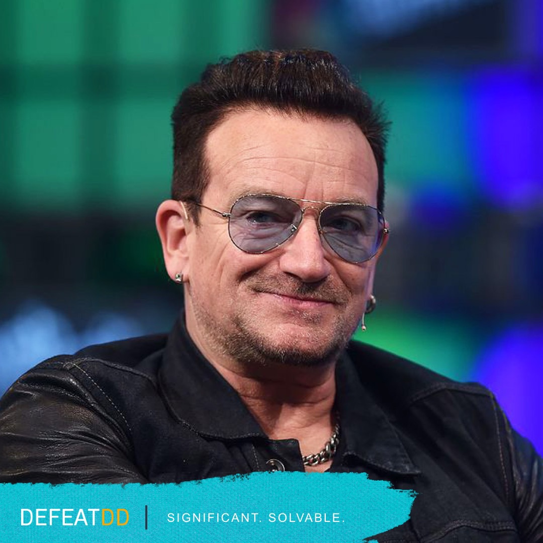 A man with short dark hair, wearing dark sunglasses and a black leather jacket, smiles at the camera. The background is a blurred mix of green and purple. The bottom-left corner has the text "DEFEATDD" and "Significant. Solvable.