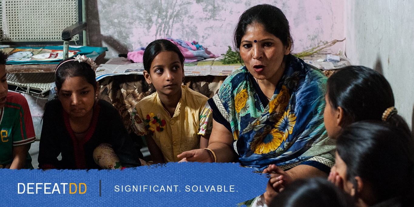 A woman wearing a blue sari with a floral pattern is speaking to a group of children, who are attentively listening. The setting appears to be a modest room with simple decor. Text at the bottom reads, "DEFEATDD. SIGNIFICANT. SOLVABLE.