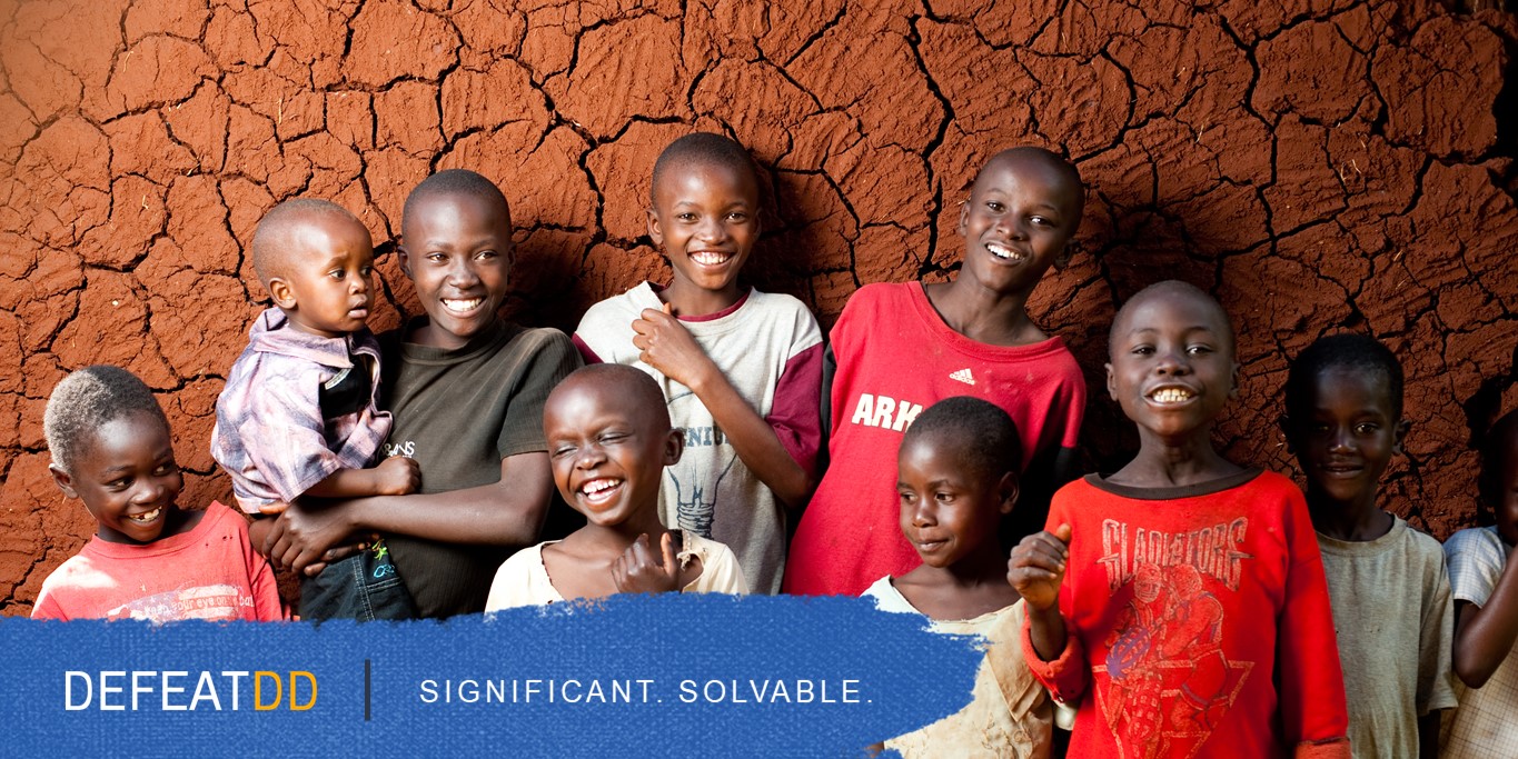 A group of nine smiling children stand together in front of a cracked, dry ground backdrop. A blue banner at the bottom reads "DEFEAT DD - Significant. Solvable.