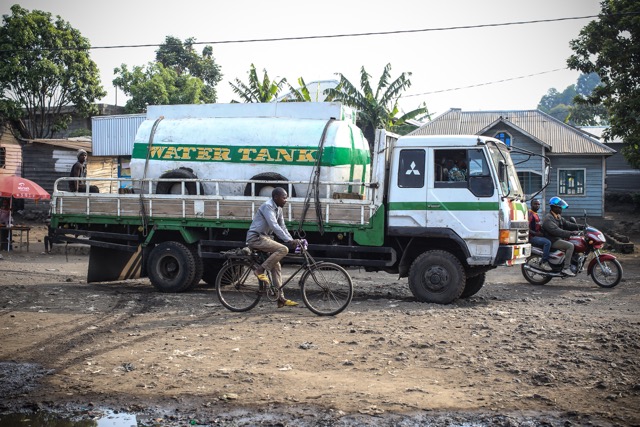 A white truck with a water tank parked on a dirt road. A man on a bicycle rides by in the foreground, while another person on a motorcycle is nearby. Houses and trees are in the background.