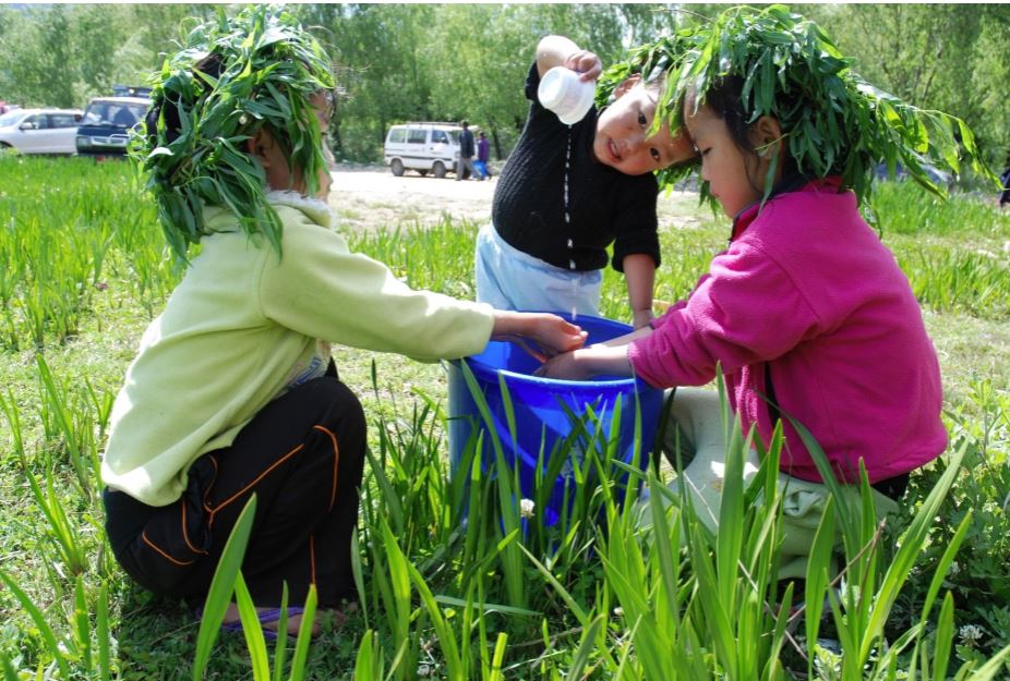 Three children wearing leafy headbands play with water in a blue tub on a grassy field. One pours water while the other two splash their hands, surrounded by greenery and parked vehicles in the background.