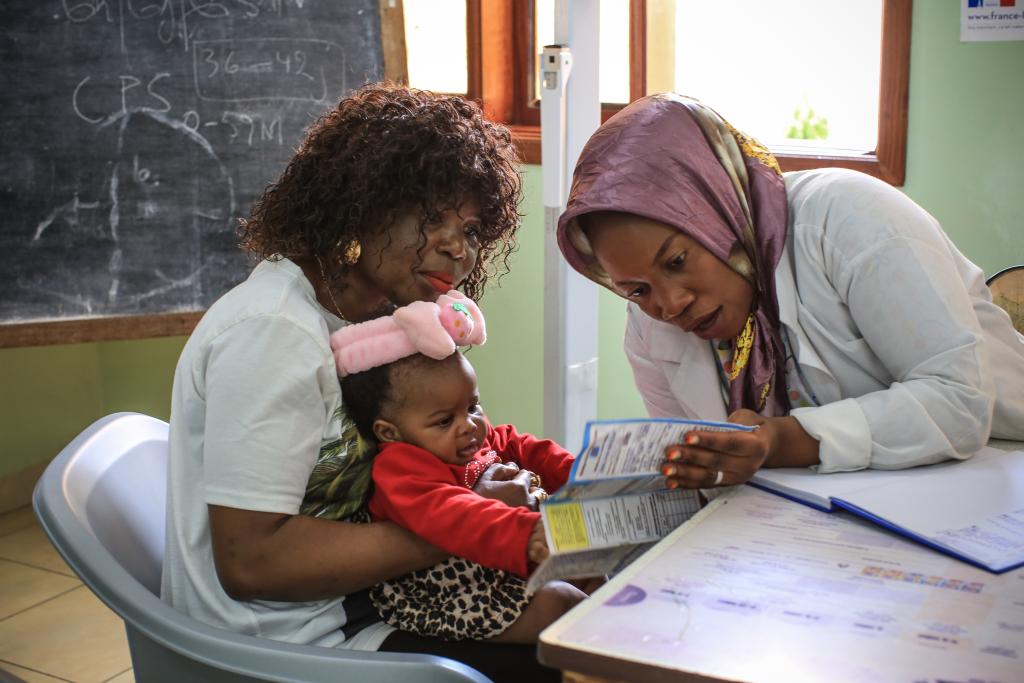 A healthcare worker in a white coat and headscarf shows a brochure to a woman holding a baby. The baby is wearing a pink headband with a stuffed animal on it. They are seated in a room with a blackboard and window in the background.