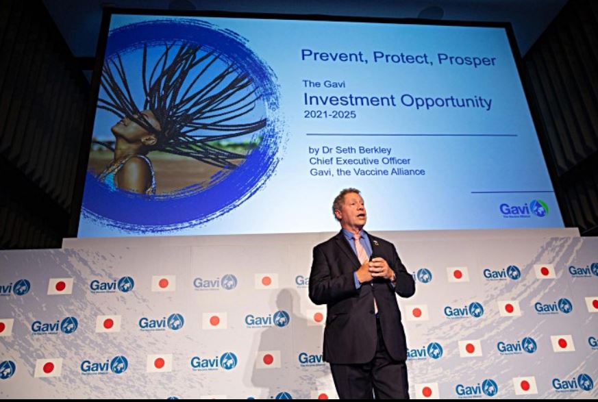 A man stands on stage in front of a large screen displaying a presentation titled "The Gavi Investment Opportunity 2021-2025" with the subtitle "Prevent, Protect, Prosper." The background features an image of a child with long braids flying in the air.