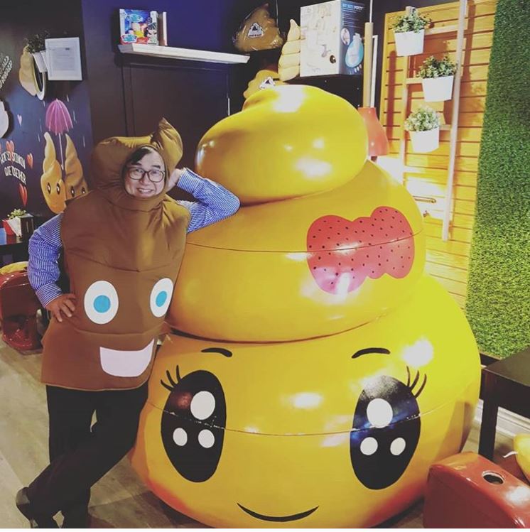 A person in a poop emoji costume smiles and leans on a giant yellow poop emoji statue with cartoon eyes and a red bow. The background features decorative elements and plants.