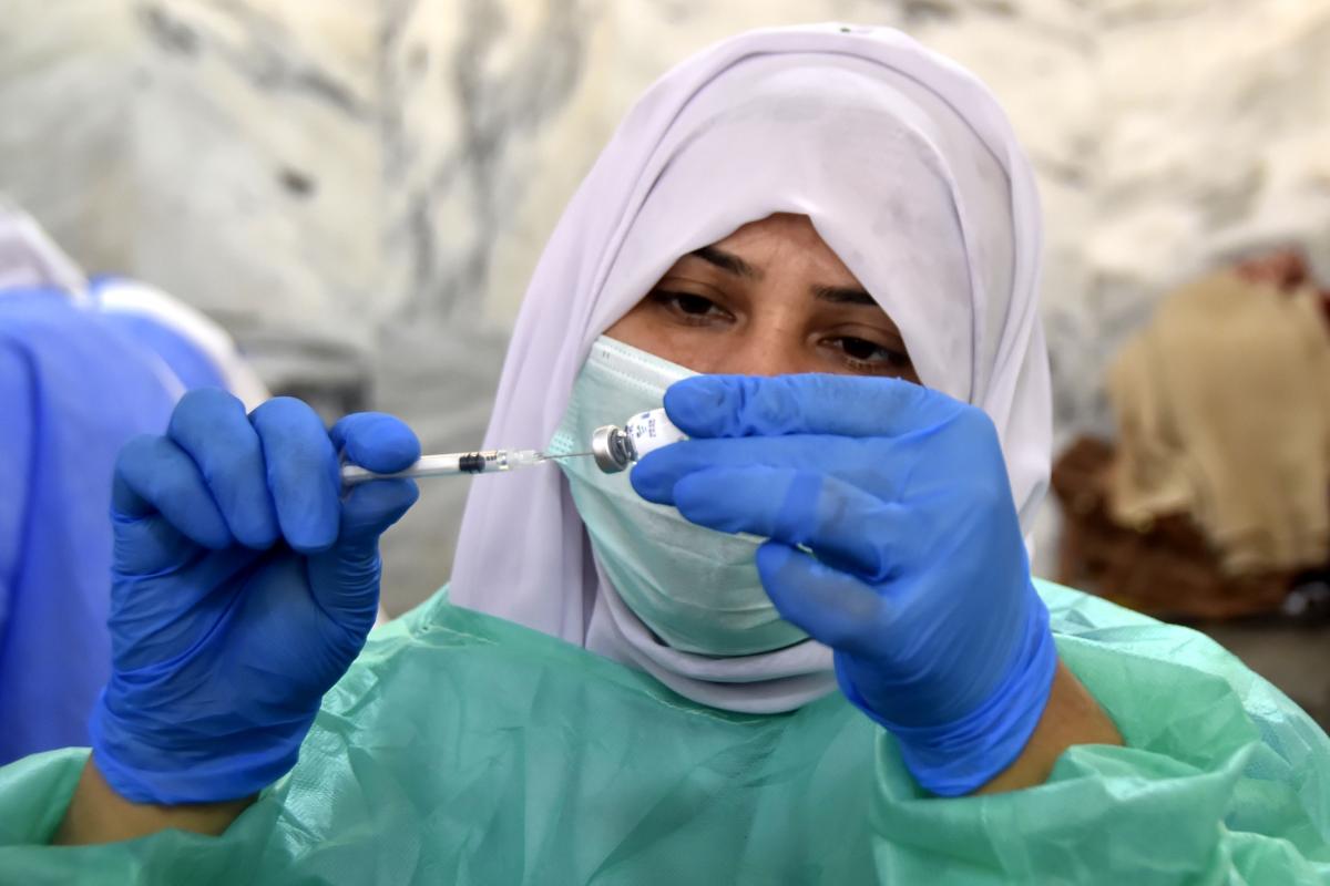 A healthcare worker wearing a face mask, headscarf, and green protective gown is preparing a syringe with a vaccine. She is holding the syringe in one hand and the vial in the other, with blue gloves on both hands. Background is out of focus.