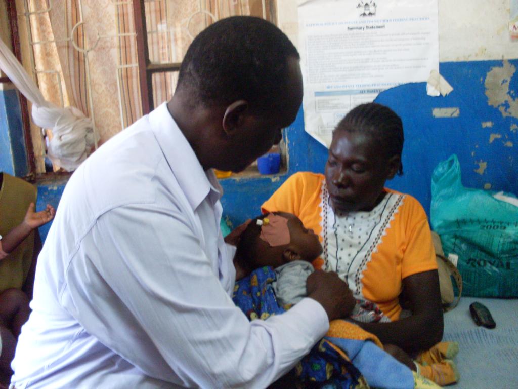 A man, wearing a white shirt, and a woman, wearing a yellow and white dress, are in a room. The man is holding a baby wrapped in a blue and orange blanket, while the woman looks on. A wall with a poster and a window with curtains are in the background.