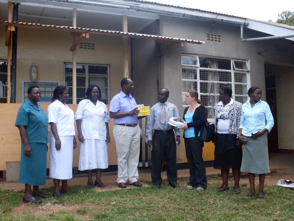 A group of seven people stands outside a building. One person is presenting a yellow booklet to another. The individuals wear a mix of professional attire, including white coats, dresses, and business casual outfits. The background shows a building with windows and a corrugated roof.