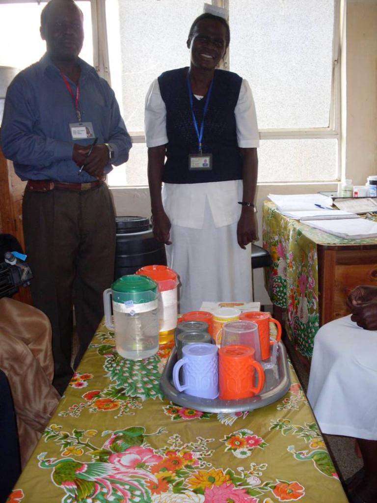 Two people are standing beside a table covered with a floral tablecloth. On the table are a silver tray with several colorful mugs and a jug of water. They are in a brightly lit room with a large window in the background.