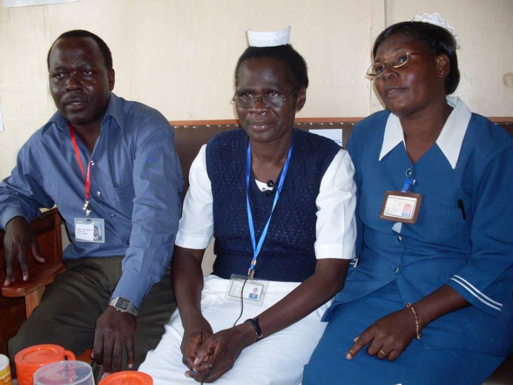 Three healthcare workers sit together, two women wearing nurse uniforms and one man in a shirt and tie. They have identification badges and are seated indoors, smiling slightly at the camera.